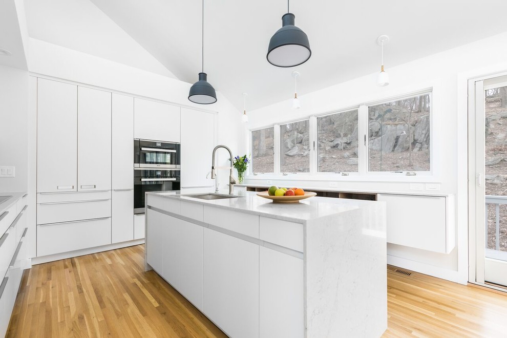 Mid-sized minimalist galley medium tone wood floor and brown floor eat-in kitchen photo in New York with a drop-in sink, white cabinets, quartz countertops, white backsplash, glass sheet backsplash, stainless steel appliances and an island