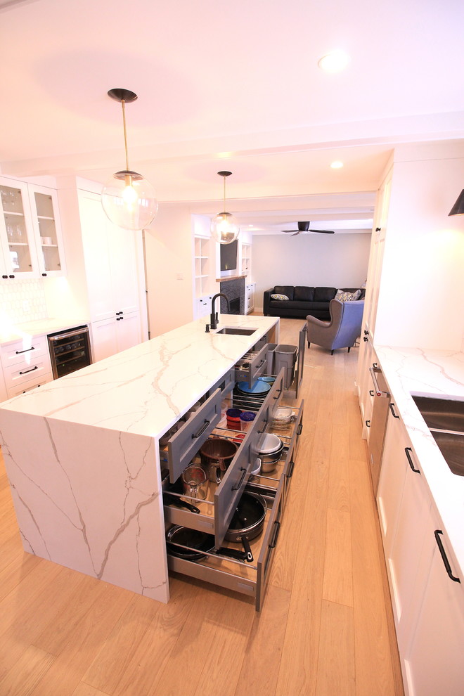 Example of a large trendy galley light wood floor enclosed kitchen design in Other with an undermount sink, shaker cabinets, white cabinets, quartz countertops, white backsplash, stone tile backsplash, stainless steel appliances and an island