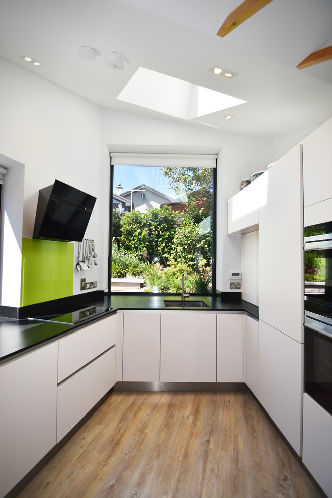 Пример оригинального дизайна: п-образная кухня среднего размера в современном стиле с плоскими фасадами, белыми фасадами, столешницей из кварцевого агломерата, полом из винила, черной столешницей, одинарной мойкой и зеленым фартуком без острова