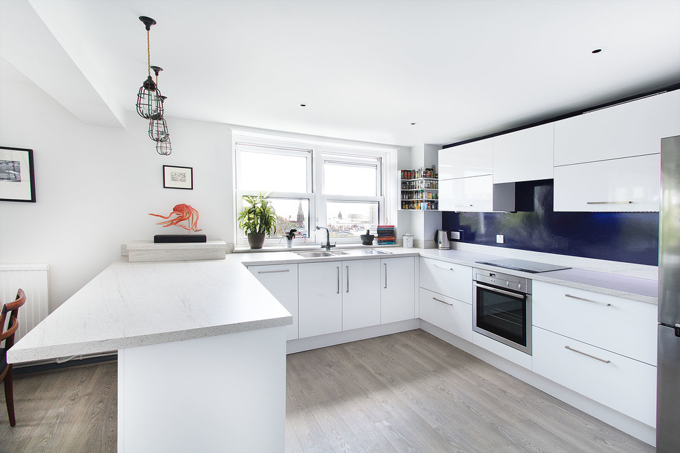 Example of a mid-sized trendy u-shaped light wood floor open concept kitchen design in Sussex with a drop-in sink, flat-panel cabinets, white cabinets, laminate countertops, blue backsplash, stainless steel appliances and a peninsula