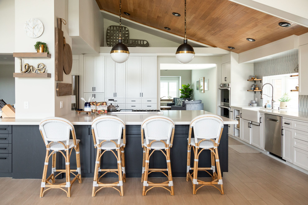 Example of a large beach style l-shaped porcelain tile and brown floor kitchen design in San Diego with a farmhouse sink, shaker cabinets, quartz countertops, ceramic backsplash, stainless steel appliances, a peninsula, white countertops, white cabinets and gray backsplash