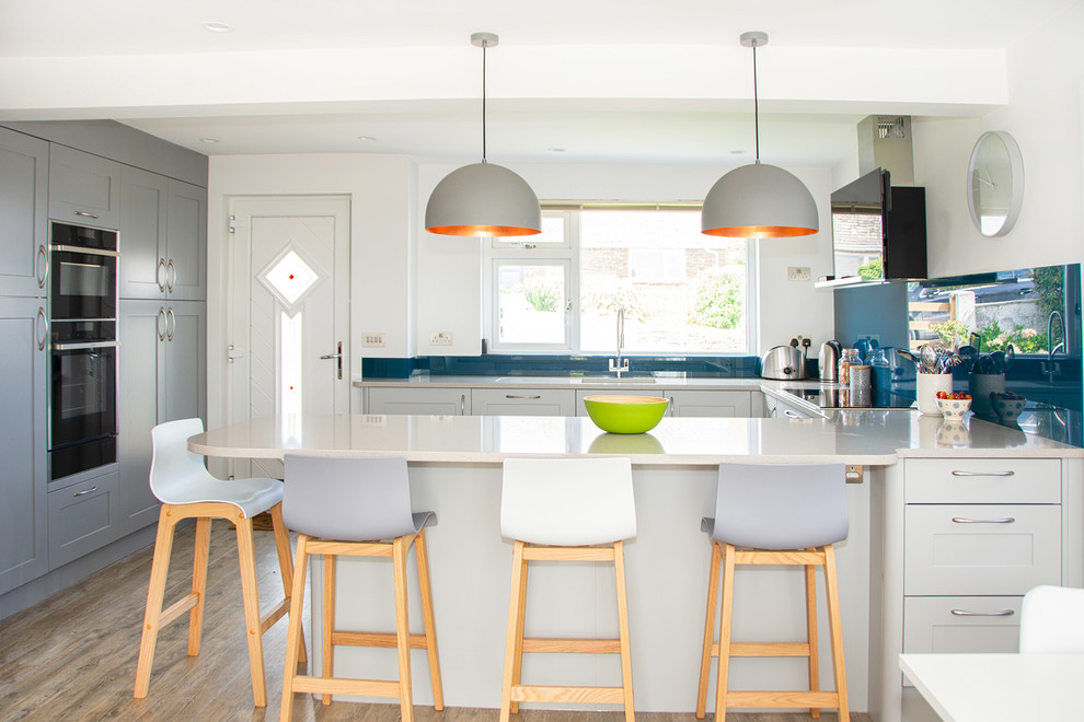 Photo of a medium sized contemporary u-shaped open plan kitchen in Cornwall with a submerged sink, recessed-panel cabinets, grey cabinets, blue splashback, stainless steel appliances, a breakfast bar, brown floors, glass sheet splashback and white worktops.