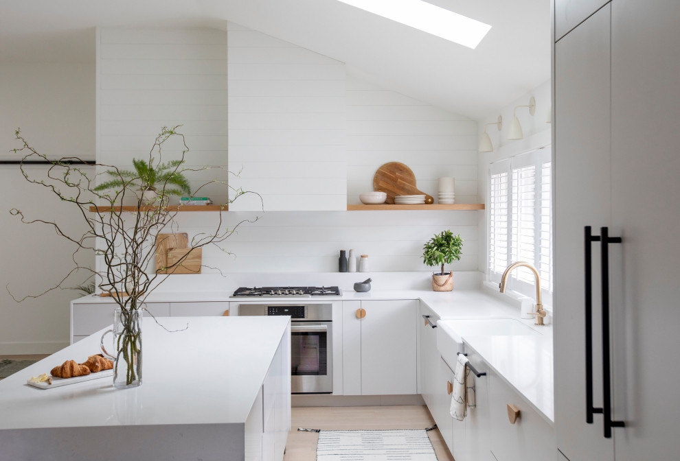 Photo of a large modern l-shaped open plan kitchen with a belfast sink, flat-panel cabinets, grey cabinets, quartz worktops, white splashback, tonge and groove splashback, integrated appliances, light hardwood flooring, an island, white worktops and a vaulted ceiling.