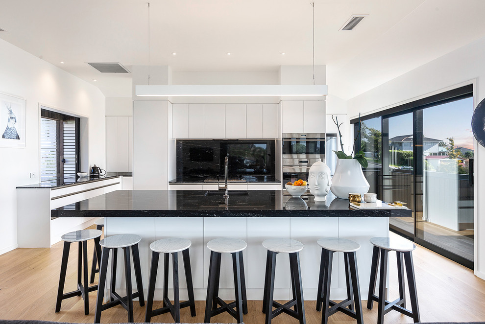 Example of a large trendy galley light wood floor and beige floor open concept kitchen design in Auckland with an undermount sink, flat-panel cabinets, granite countertops, black backsplash, stone slab backsplash, stainless steel appliances, an island and black countertops