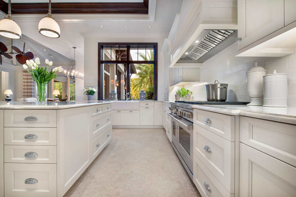Example of a large island style l-shaped limestone floor and beige floor eat-in kitchen design in Miami with a farmhouse sink, shaker cabinets, white cabinets, marble countertops, white backsplash, stone slab backsplash, paneled appliances and an island