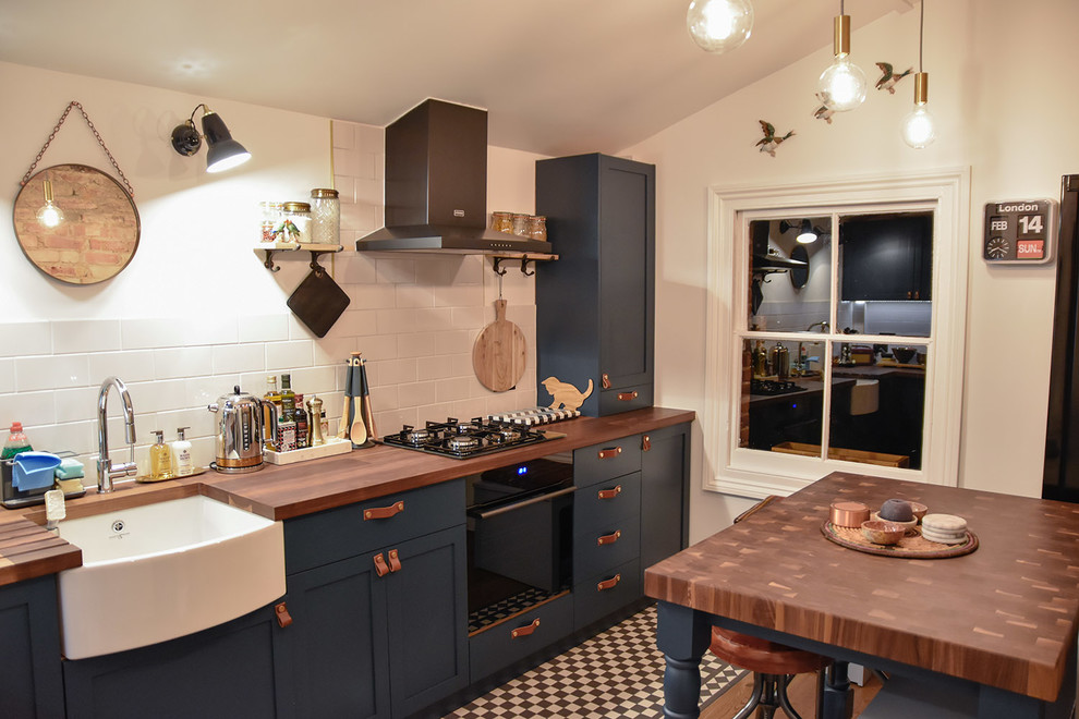 Modern inredning av ett mellanstort kök, med en rustik diskho, skåp i shakerstil, blå skåp, träbänkskiva, vitt stänkskydd, stänkskydd i keramik, svarta vitvaror, klinkergolv i keramik och en halv köksö