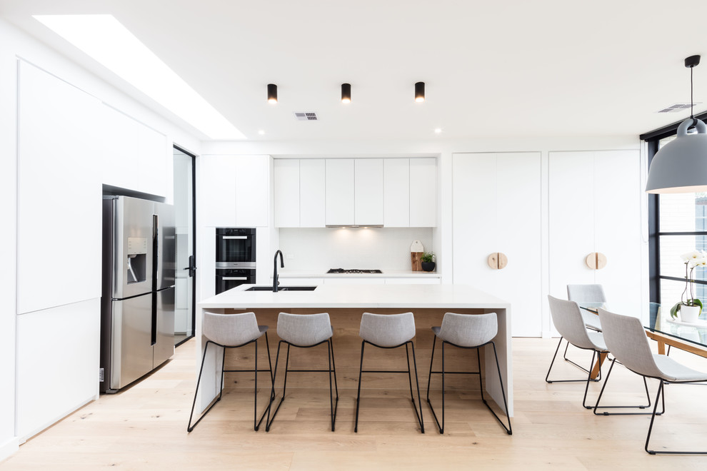 Moderne Wohnküche in L-Form mit Doppelwaschbecken, flächenbündigen Schrankfronten, Küchenrückwand in Weiß, Küchengeräten aus Edelstahl, hellem Holzboden, Kücheninsel, beigem Boden und weißer Arbeitsplatte in Melbourne