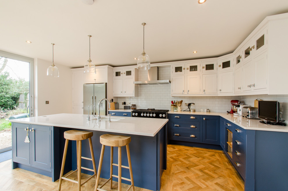 Large country l-shaped kitchen/diner in Kent with an integrated sink, shaker cabinets, blue cabinets, quartz worktops, metro tiled splashback, coloured appliances, medium hardwood flooring and an island.