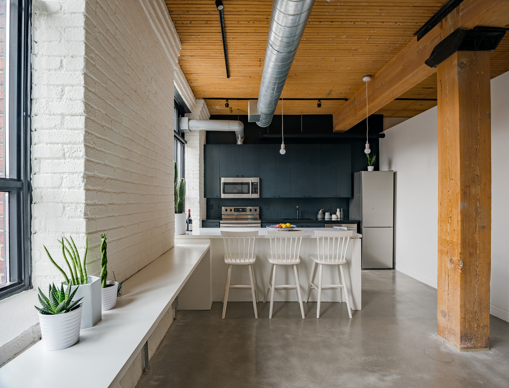 Foto på ett industriellt kök, med släta luckor, svarta skåp, svart stänkskydd, rostfria vitvaror, betonggolv, en halv köksö och grått golv