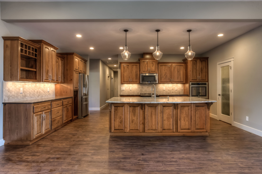 Mountain style brown floor open concept kitchen photo in Portland with a farmhouse sink, medium tone wood cabinets, granite countertops, beige backsplash, stainless steel appliances, an island, multicolored countertops and raised-panel cabinets
