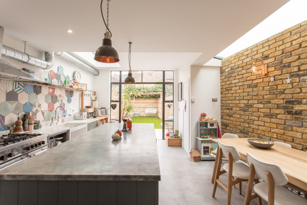 This is an example of an industrial galley kitchen in London with a belfast sink, grey cabinets, multi-coloured splashback, cement tile splashback, stainless steel appliances, an island, grey floors and grey worktops.