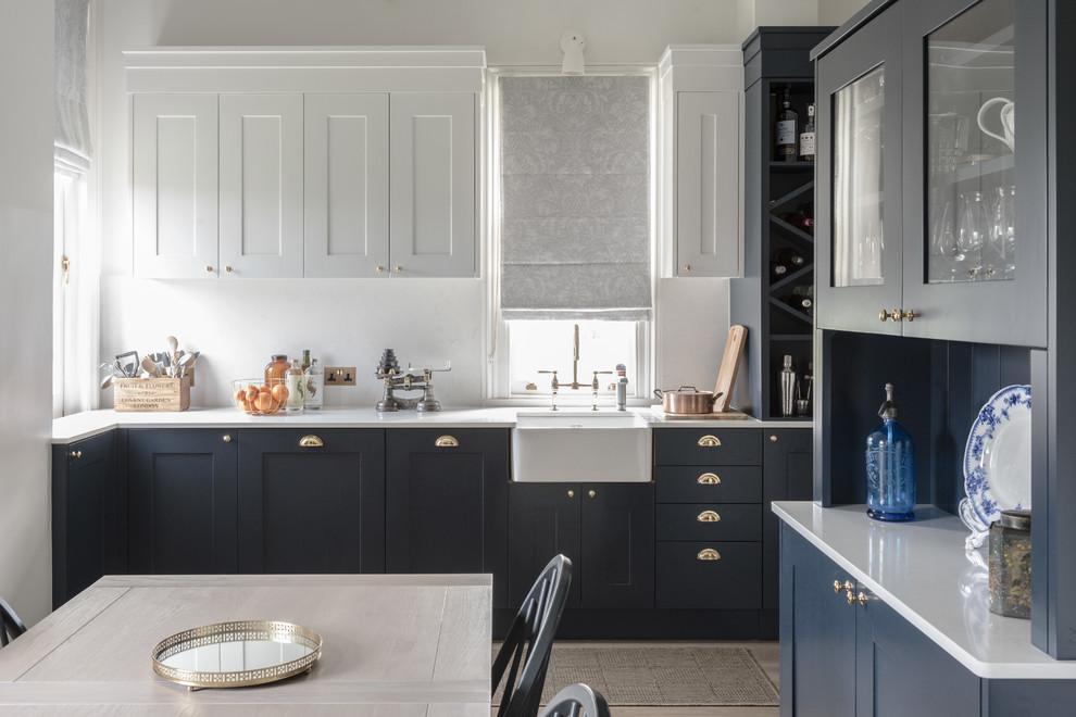 Contemporary l-shaped kitchen/diner in London with a belfast sink, shaker cabinets, blue cabinets, composite countertops, white splashback, marble splashback, integrated appliances and light hardwood flooring.