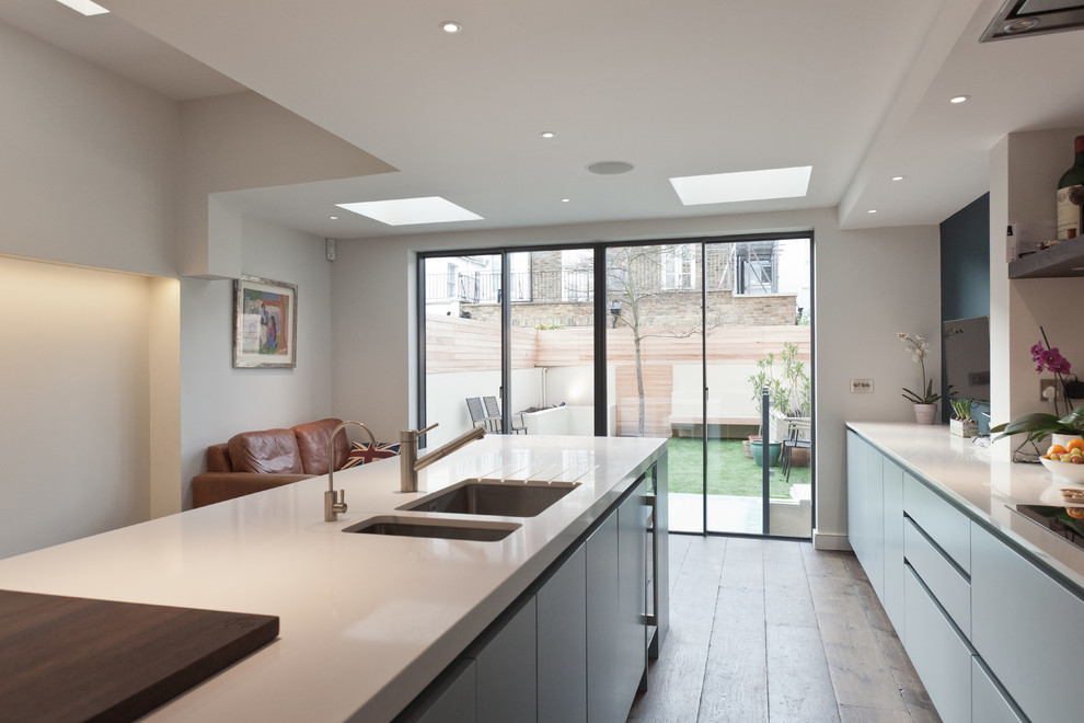 Photo of a medium sized contemporary single-wall open plan kitchen in London with flat-panel cabinets, white splashback, dark hardwood flooring, an island, an integrated sink, grey cabinets, composite countertops, glass sheet splashback, integrated appliances and white worktops.