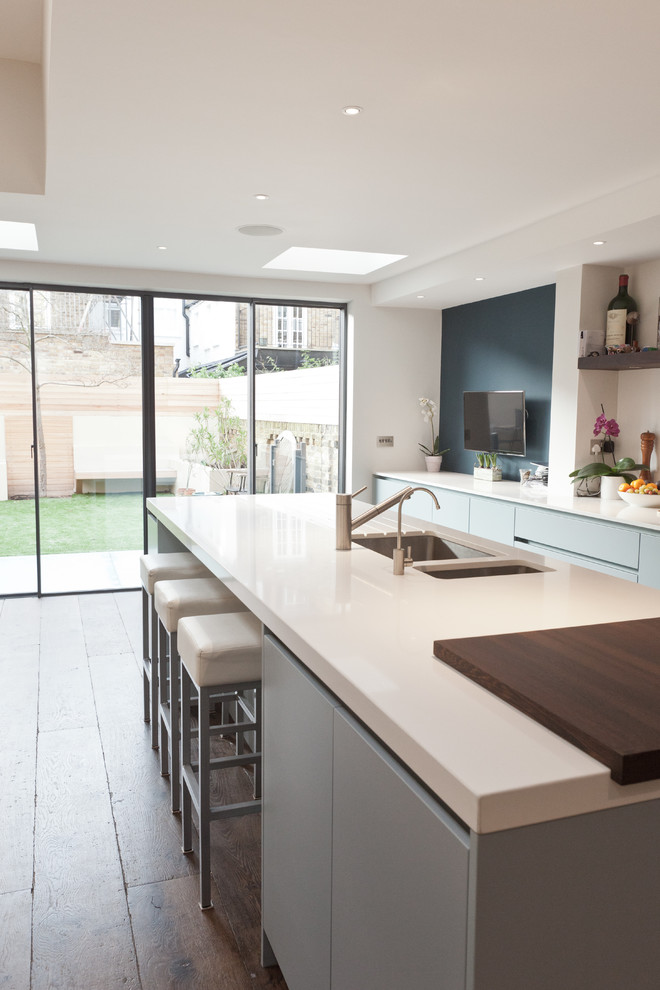 Photo of a large contemporary single-wall kitchen/diner in London with flat-panel cabinets, white splashback, dark hardwood flooring, an island, an integrated sink, grey cabinets, composite countertops, glass sheet splashback, white worktops and integrated appliances.