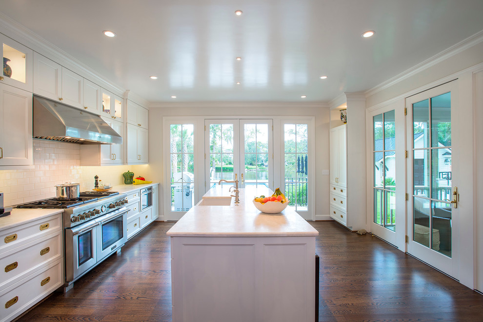 Inspiration for a large classic single-wall kitchen/diner in Charleston with a belfast sink, recessed-panel cabinets, white cabinets, engineered stone countertops, white splashback, ceramic splashback, stainless steel appliances, medium hardwood flooring, an island and brown floors.
