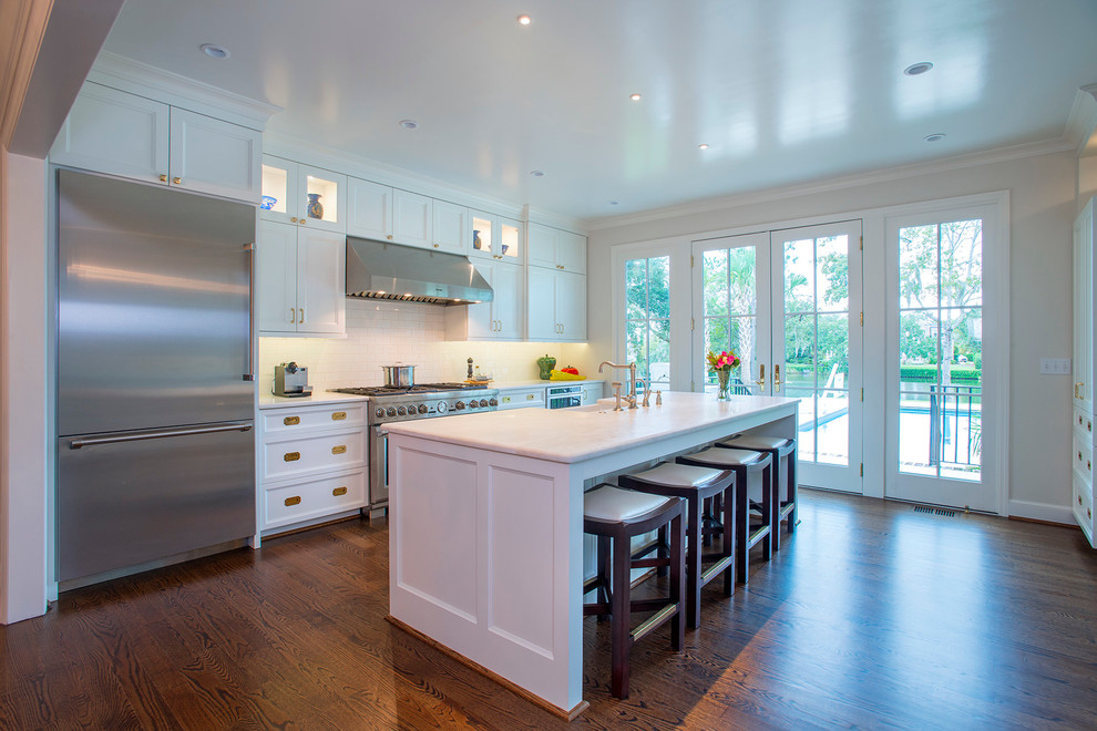 This is an example of a large classic single-wall kitchen/diner in Charleston with a belfast sink, recessed-panel cabinets, white cabinets, engineered stone countertops, white splashback, ceramic splashback, stainless steel appliances, medium hardwood flooring, an island and brown floors.