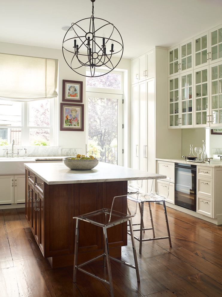 Klassische Küche mit weißen Schränken, Marmor-Arbeitsplatte, Kücheninsel, Landhausspüle, Glasfronten und braunem Holzboden in New York