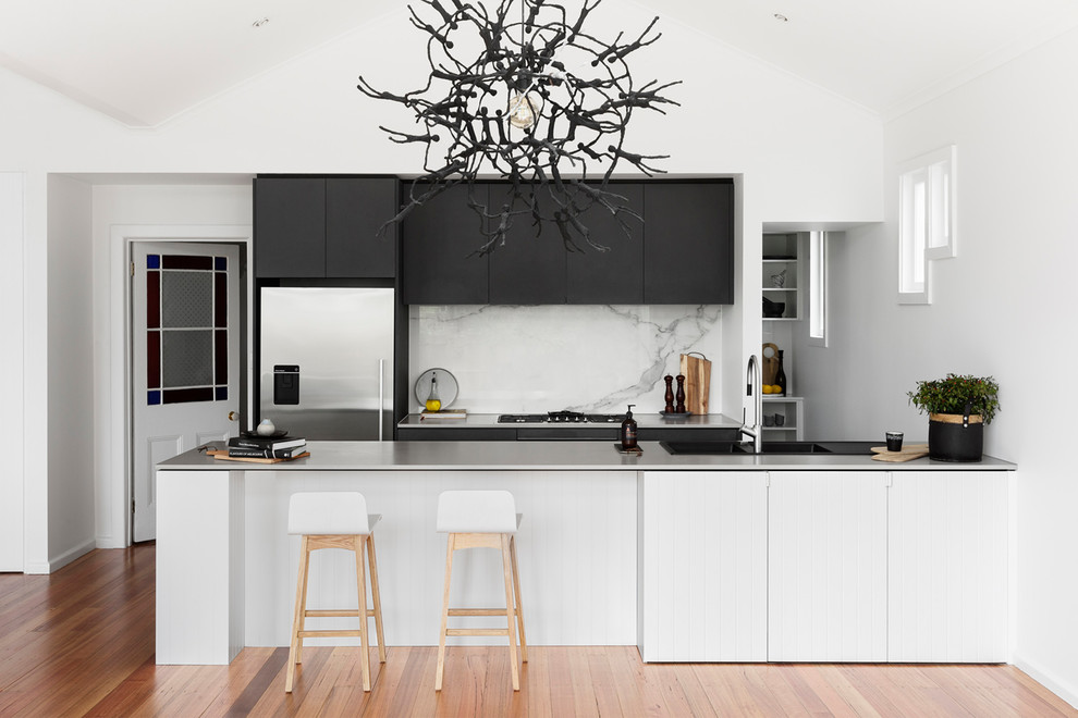 Bild på ett mellanstort funkis vit vitt kök, med en integrerad diskho, släta luckor, bänkskiva i rostfritt stål, vitt stänkskydd, rostfria vitvaror, mellanmörkt trägolv, en halv köksö, stänkskydd i marmor och brunt golv