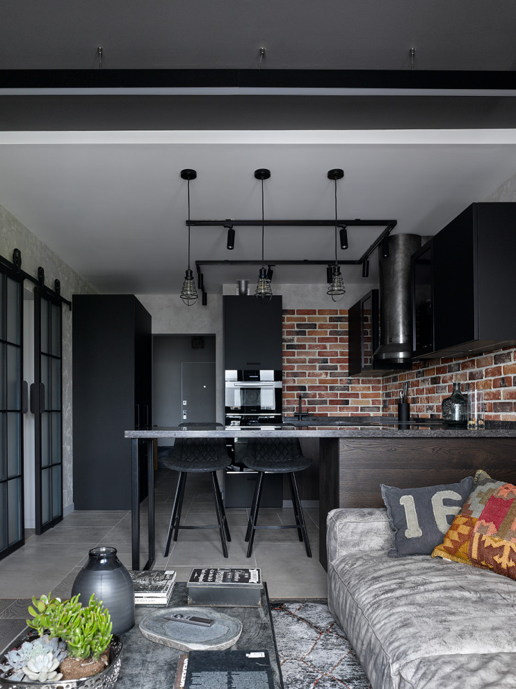 На фото: п-образная кухня-гостиная среднего размера в стиле лофт с плоскими фасадами, черными фасадами, коричневым фартуком, фартуком из кирпича, черной техникой, полуостровом, серым полом и черной столешницей