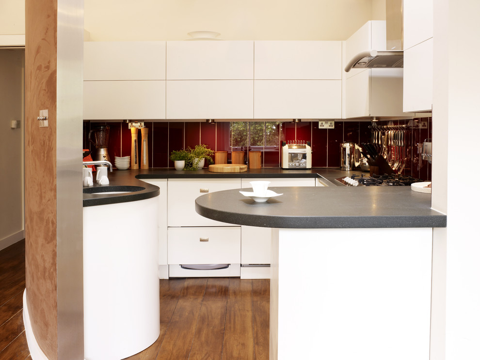 Mid-sized trendy u-shaped medium tone wood floor open concept kitchen photo in London with flat-panel cabinets, white cabinets, red backsplash, a peninsula, a drop-in sink, wood countertops and paneled appliances