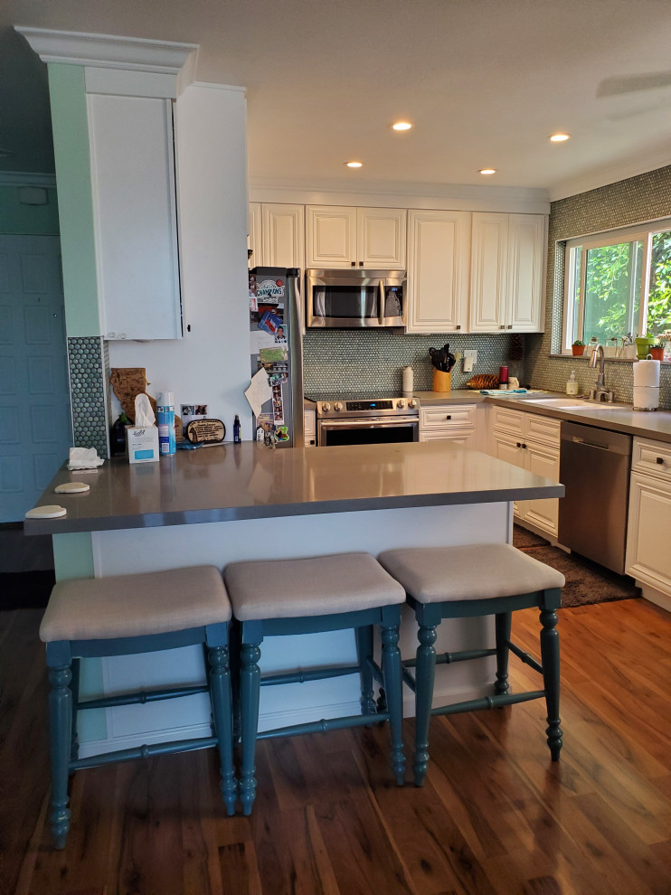 Bild på ett litet maritimt grå grått kök, med en undermonterad diskho, luckor med upphöjd panel, vita skåp, bänkskiva i kvartsit, grönt stänkskydd, stänkskydd i glaskakel, rostfria vitvaror, mellanmörkt trägolv, en köksö och brunt golv