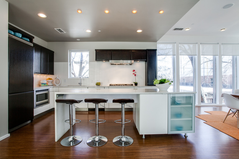 Moderne Wohnküche mit dunklen Holzschränken, Küchenrückwand in Weiß, Elektrogeräten mit Frontblende, braunem Holzboden und Kücheninsel in Dallas