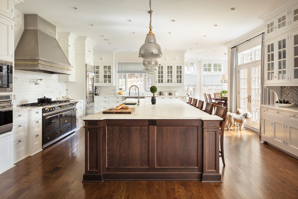Example of a classic u-shaped medium tone wood floor and brown floor eat-in kitchen design in New York with a farmhouse sink, white cabinets, white backsplash, subway tile backsplash, stainless steel appliances, an island, white countertops and recessed-panel cabinets