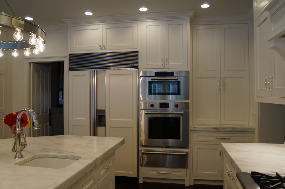 Klassisk inredning av ett mellanstort vit vitt kök, med en undermonterad diskho, luckor med infälld panel, vita skåp, marmorbänkskiva, vitt stänkskydd, stänkskydd i sten, rostfria vitvaror, flera köksöar, mörkt trägolv och brunt golv