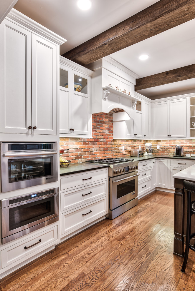 Photo of a large rural l-shaped kitchen/diner in New York with a belfast sink, beaded cabinets, white cabinets, granite worktops, brick splashback, stainless steel appliances, an island, brown floors, black worktops and painted wood flooring.