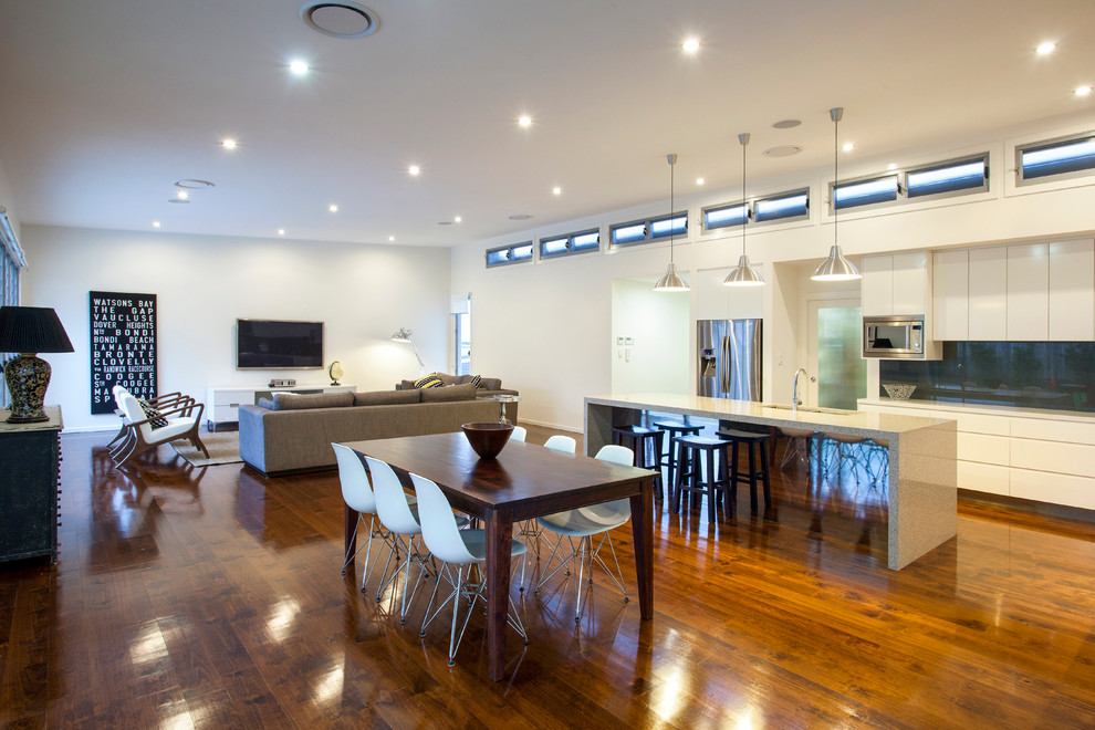 Photo of a medium sized contemporary l-shaped open plan kitchen in Brisbane with a double-bowl sink, white cabinets, composite countertops, metallic splashback, glass sheet splashback, stainless steel appliances, medium hardwood flooring and an island.