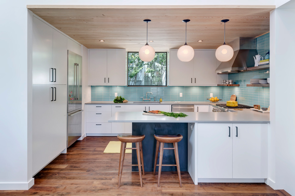 Bild på ett funkis u-kök, med en undermonterad diskho, släta luckor, vita skåp, blått stänkskydd, stänkskydd i tunnelbanekakel, rostfria vitvaror, mörkt trägolv och en halv köksö