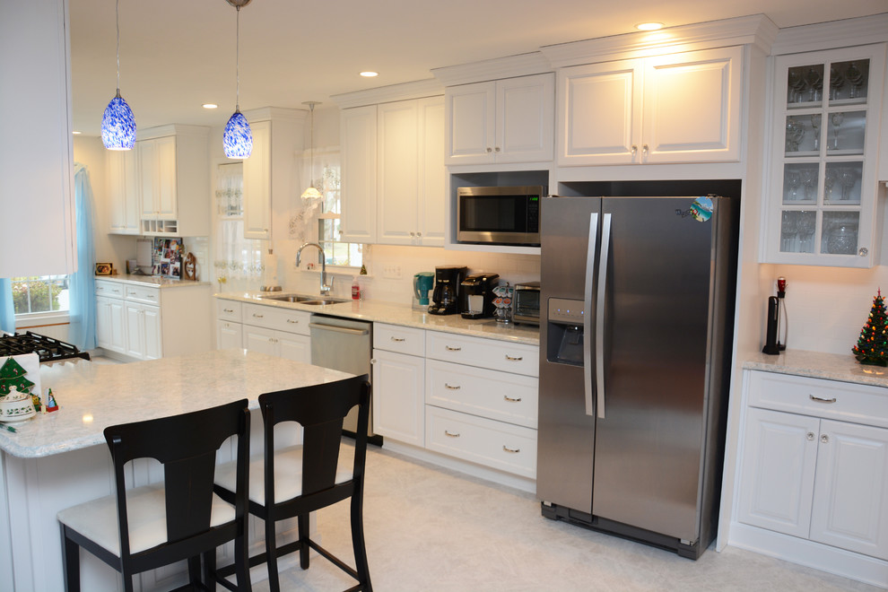 This is an example of a large classic single-wall open plan kitchen in DC Metro with a double-bowl sink, raised-panel cabinets, white cabinets, engineered stone countertops, white splashback, ceramic splashback, stainless steel appliances, a breakfast bar, white floors and white worktops.