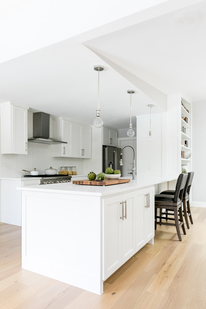 Traditional galley kitchen in Austin with shaker cabinets, white cabinets, white splashback, stainless steel appliances, light hardwood flooring, a breakfast bar, beige floors and white worktops.