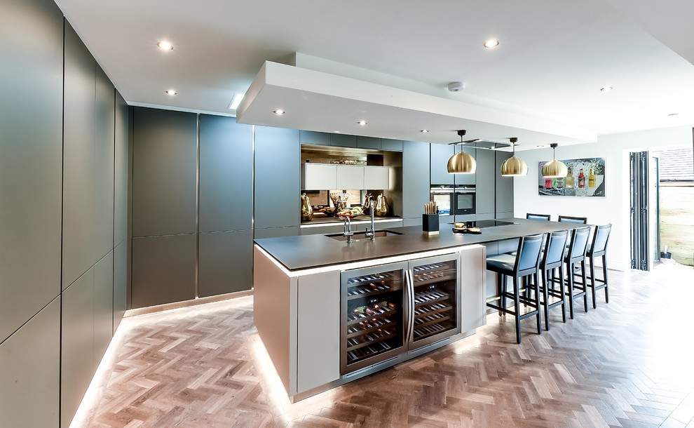 Large contemporary l-shaped kitchen in Hertfordshire with a submerged sink, flat-panel cabinets, grey cabinets, mirror splashback, stainless steel appliances, light hardwood flooring, an island, brown floors and black worktops.