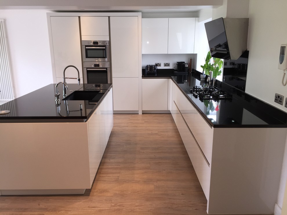 Inspiration for a medium sized contemporary l-shaped kitchen/diner in Hertfordshire with a submerged sink, flat-panel cabinets, white cabinets, granite worktops, black splashback, glass sheet splashback, stainless steel appliances, medium hardwood flooring and an island.