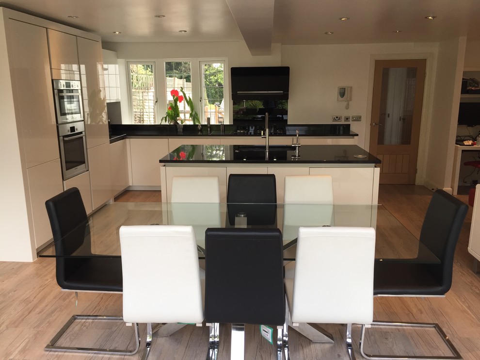 Mid-sized trendy l-shaped medium tone wood floor eat-in kitchen photo in Hertfordshire with an undermount sink, flat-panel cabinets, white cabinets, granite countertops, black backsplash, glass sheet backsplash, stainless steel appliances and an island