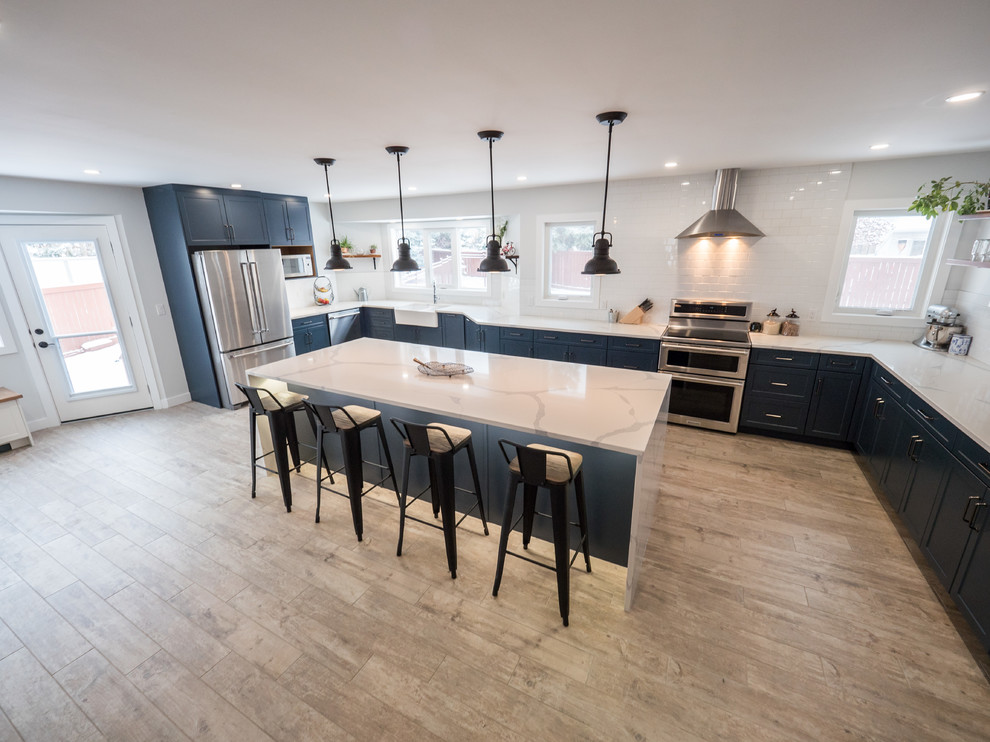 Inspiration for a large contemporary u-shaped open plan kitchen in Edmonton with an island, a belfast sink, shaker cabinets, blue cabinets, marble worktops, white splashback, metro tiled splashback, stainless steel appliances, medium hardwood flooring, brown floors and white worktops.
