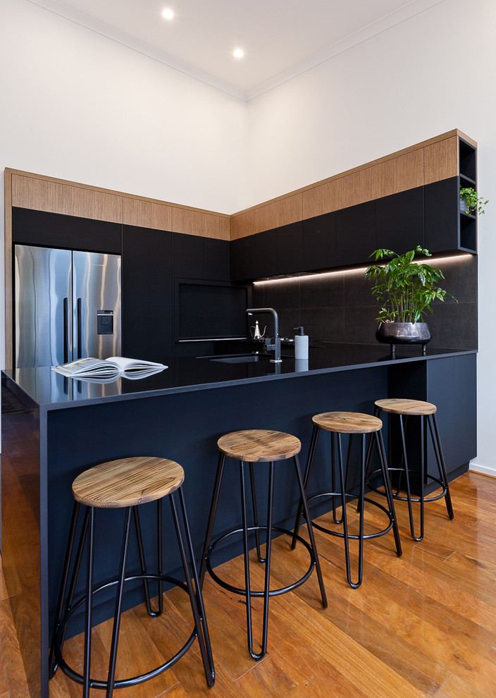 This is an example of a medium sized urban u-shaped open plan kitchen in Perth with a submerged sink, black cabinets, engineered stone countertops, black splashback, porcelain splashback, black appliances, medium hardwood flooring, a breakfast bar and black worktops.