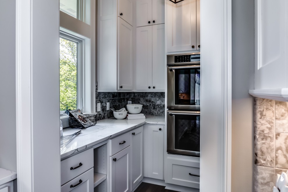 Design ideas for a small traditional galley kitchen pantry in Minneapolis with a belfast sink, shaker cabinets, white cabinets, marble worktops, black splashback, marble splashback, integrated appliances, dark hardwood flooring, no island, brown floors and white worktops.