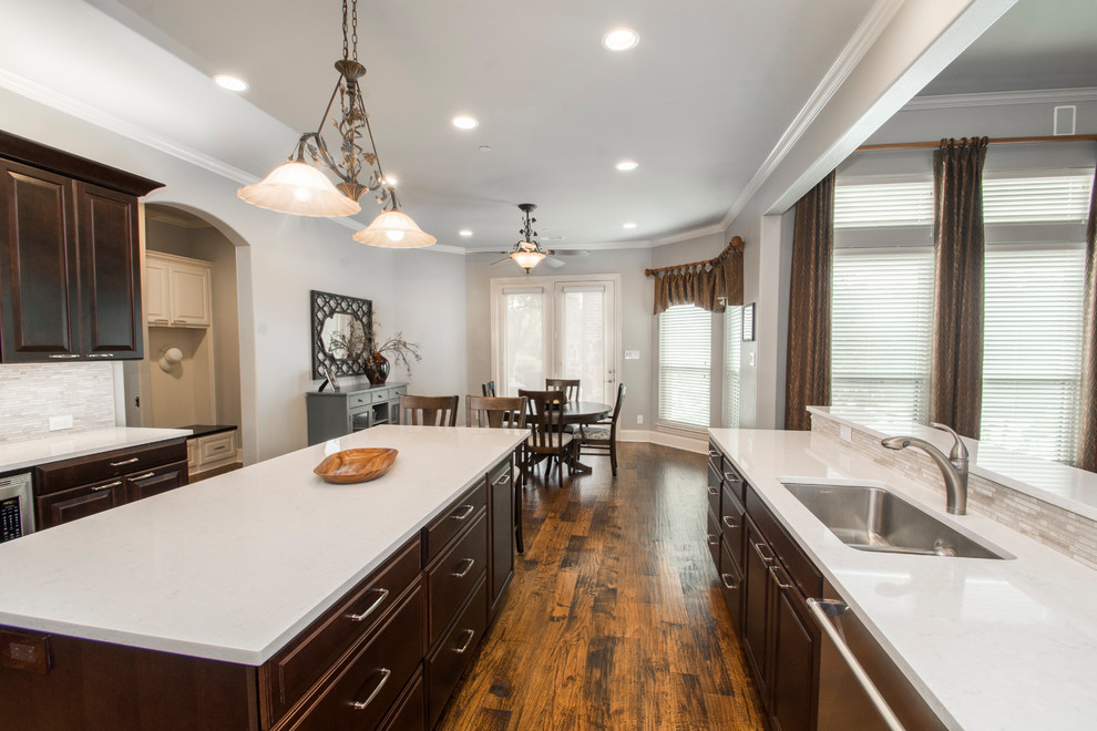 This is an example of a large classic u-shaped open plan kitchen in Dallas with a double-bowl sink, raised-panel cabinets, dark wood cabinets, engineered stone countertops, beige splashback, mosaic tiled splashback, stainless steel appliances, medium hardwood flooring, an island and brown floors.