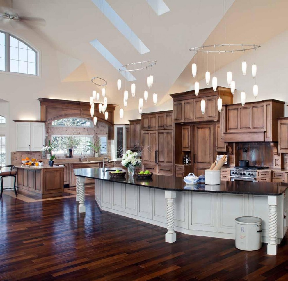 Huge trendy u-shaped dark wood floor, brown floor and vaulted ceiling open concept kitchen photo in Other with an undermount sink, raised-panel cabinets, brown cabinets, quartz countertops, brown backsplash, travertine backsplash, paneled appliances, an island and black countertops
