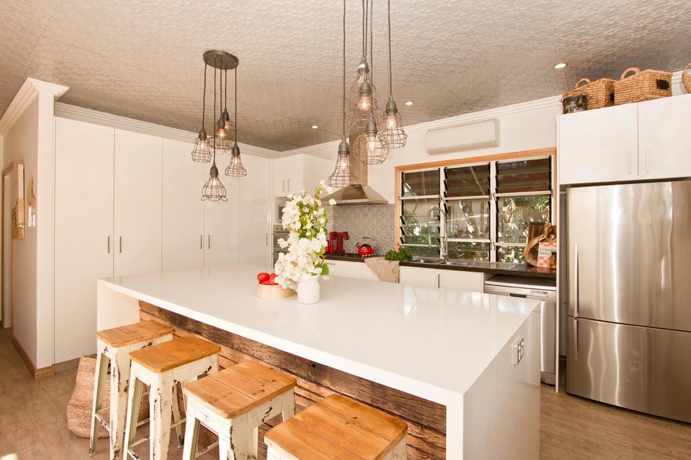 This is an example of a medium sized beach style galley kitchen in Dunedin with flat-panel cabinets, white cabinets, engineered stone countertops, stainless steel appliances, light hardwood flooring, an island, metallic splashback and metal splashback.
