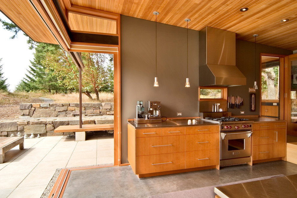 Foto på ett litet funkis linjärt kök med öppen planlösning, med släta luckor, skåp i mellenmörkt trä, bänkskiva i rostfritt stål, rostfria vitvaror, betonggolv och en köksö