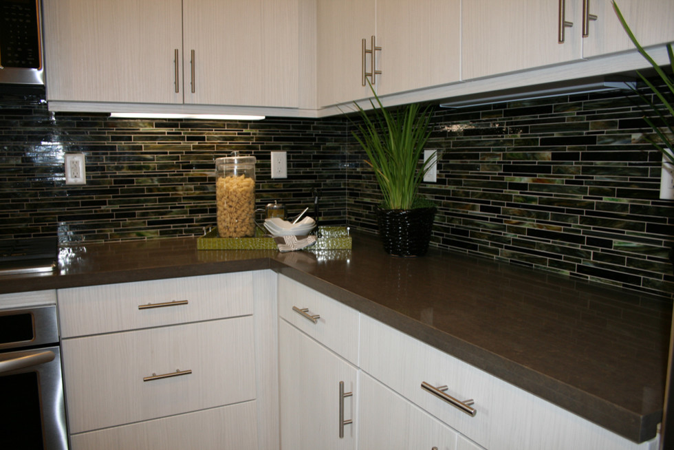 Modelo de cocina actual con armarios con paneles lisos, puertas de armario de madera clara, encimera de cuarcita, salpicadero negro y salpicadero de azulejos en listel
