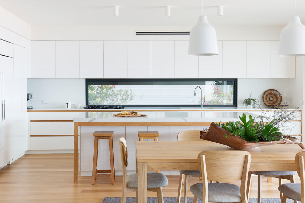 Moderne Wohnküche in L-Form mit Quarzwerkstein-Arbeitsplatte, weißer Arbeitsplatte, flächenbündigen Schrankfronten, weißen Schränken, Rückwand-Fenster, weißen Elektrogeräten, braunem Holzboden, Kücheninsel und braunem Boden in Sydney