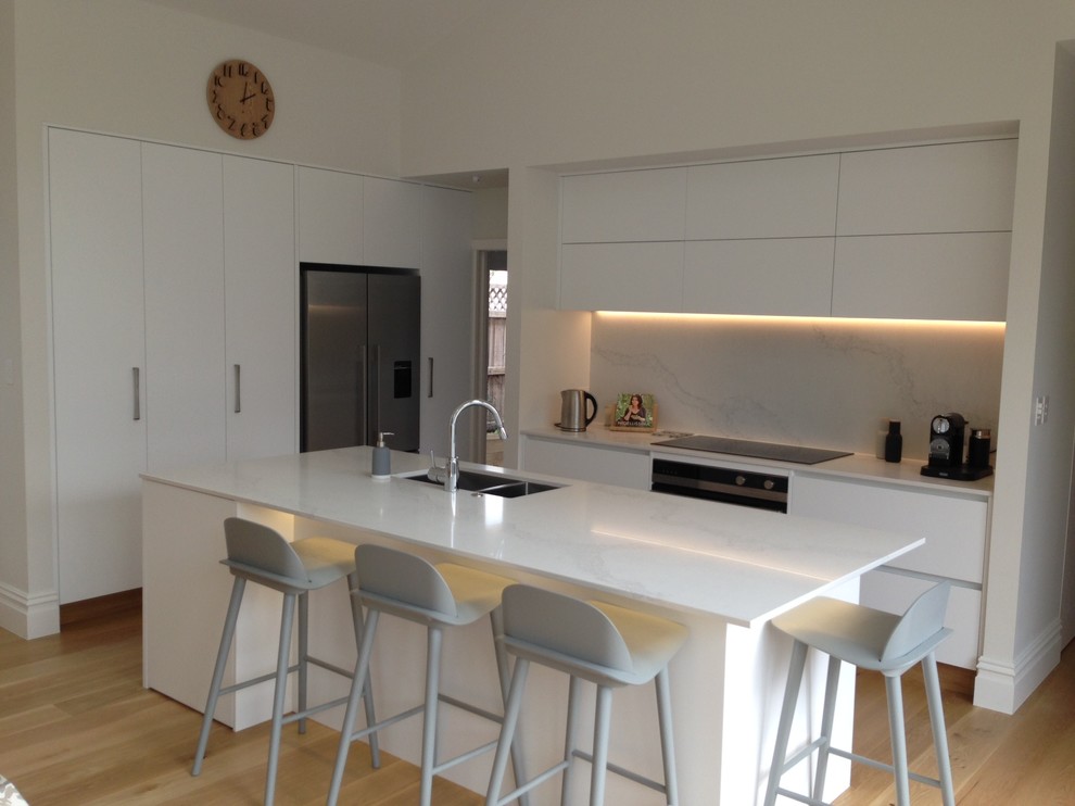 Photo of a large scandinavian l-shaped open plan kitchen in Auckland with a double-bowl sink, white cabinets, engineered stone countertops, white splashback, stone slab splashback, stainless steel appliances, light hardwood flooring and an island.