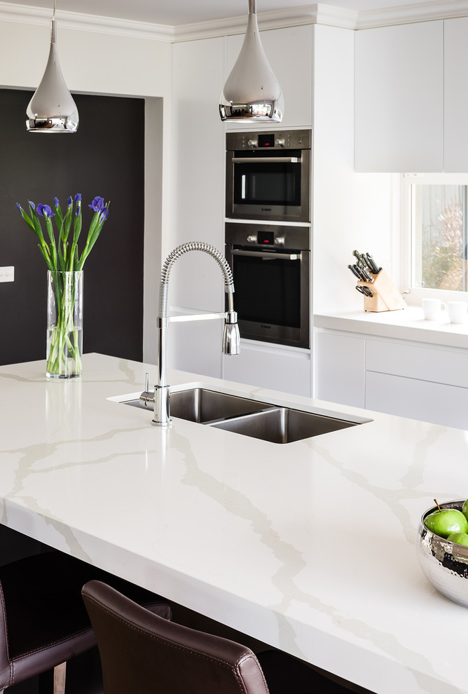Zweizeilige Moderne Wohnküche mit Unterbauwaschbecken, Schrankfronten mit vertiefter Füllung, weißen Schränken, Quarzwerkstein-Arbeitsplatte, Glasrückwand, Küchengeräten aus Edelstahl, Kücheninsel und braunem Holzboden in Sydney