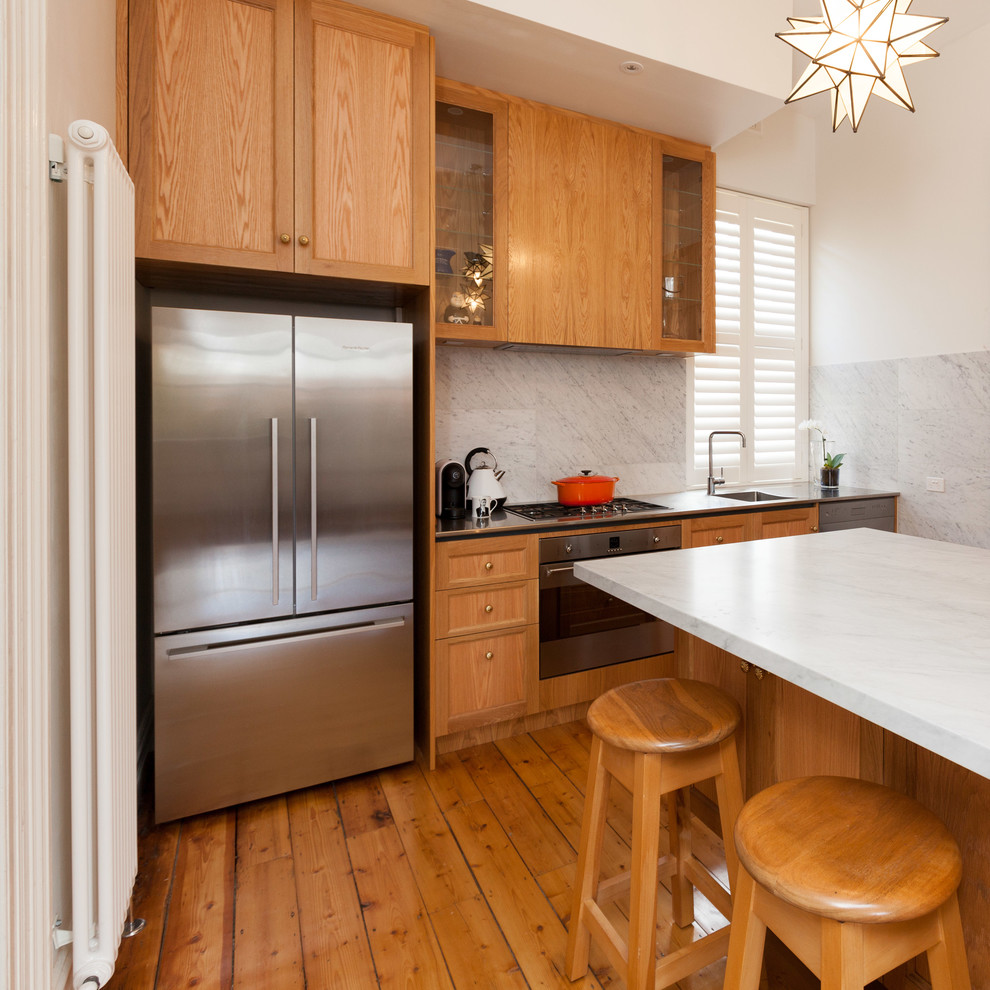 Example of a mid-sized ornate l-shaped light wood floor kitchen pantry design in Melbourne with an integrated sink, glass-front cabinets, light wood cabinets, stainless steel countertops, white backsplash, stone tile backsplash, stainless steel appliances and an island