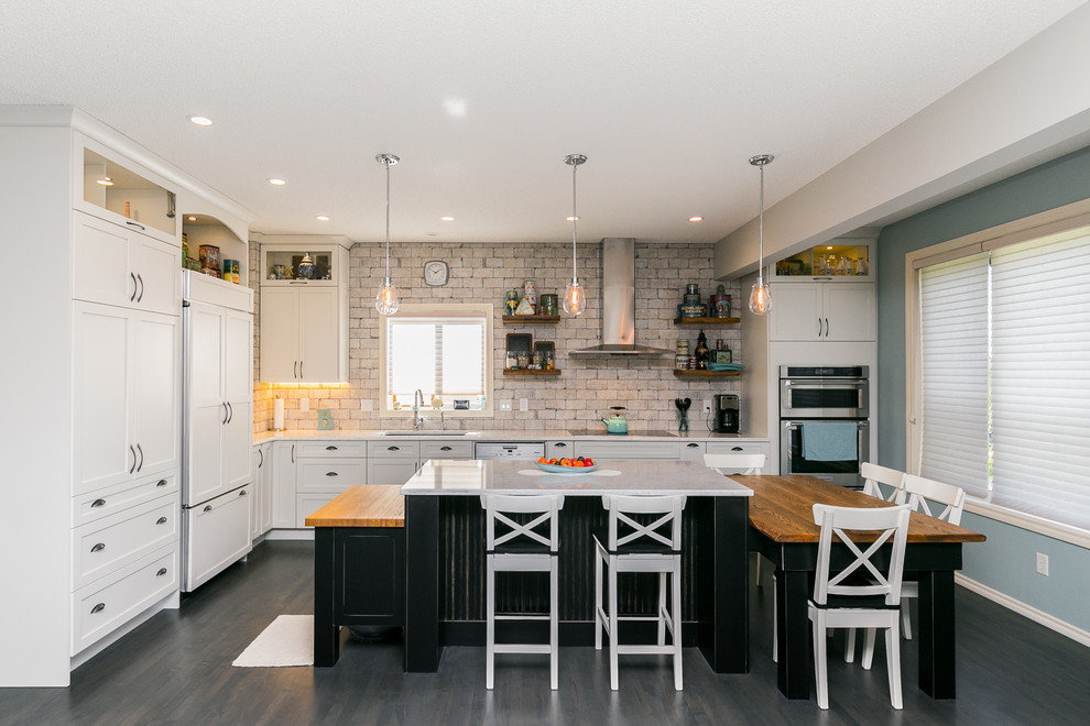 Photo of a large industrial l-shaped kitchen/diner in Calgary with shaker cabinets, white cabinets, white splashback, dark hardwood flooring, multiple islands, a submerged sink, marble worktops, brick splashback, stainless steel appliances and grey floors.