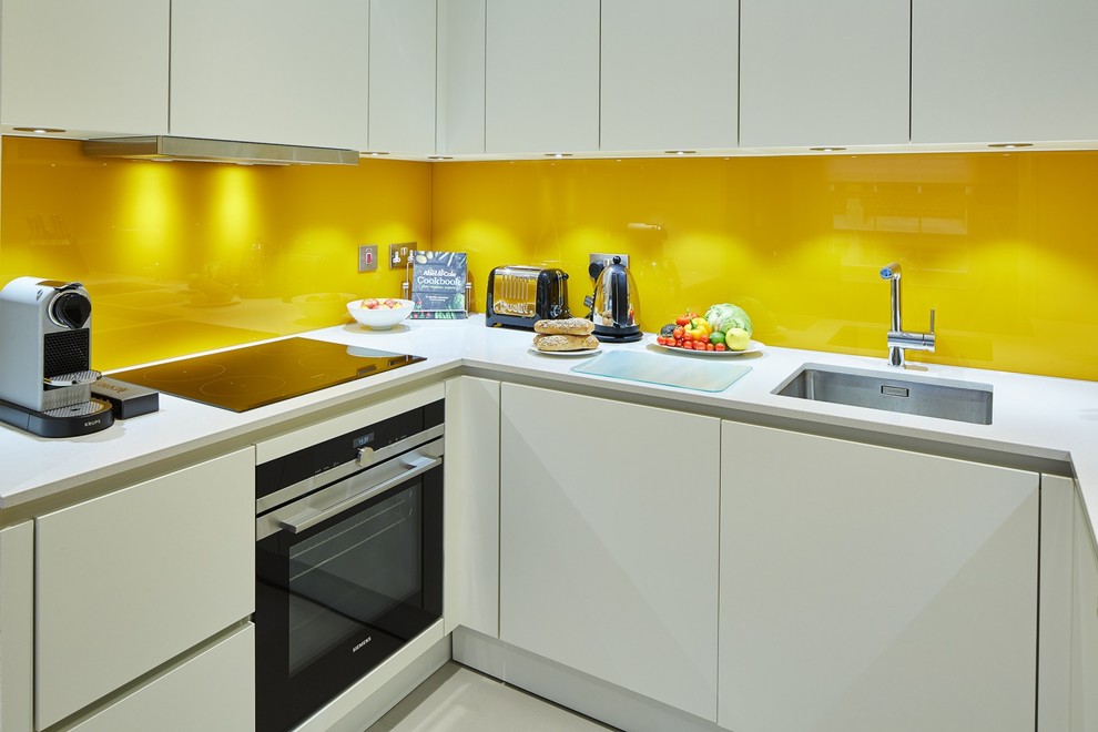 Foto på ett funkis vit kök, med en undermonterad diskho, släta luckor, vita skåp, gult stänkskydd, glaspanel som stänkskydd, rostfria vitvaror och beiget golv